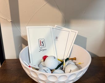 Pode incluir: Uma tigela branca em forma de bola de golfe cheia de bolas de golfe, tees e cartões de pontuação. Os cartões de pontuação têm o logotipo Sawgrass. A tigela está sobre uma superfície de madeira escura.