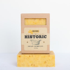 May include: A rectangular bar of yellow speckled soap sits beneath a matching soap bar encased in a brown cardboard box. The label features the text Gold Rush, Historic Soap Company, and 4.5-5oz, along with a graphic of a vintage bathtub.