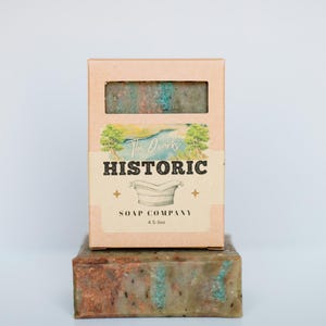 May include: A rectangular bar of mottled green and copper-toned soap sits beneath a matching cardboard box. The label features a landscape illustration, the text The Ozarks HISTORIC SOAP COMPANY, a bathtub icon, and the weight 4.5-5oz.
