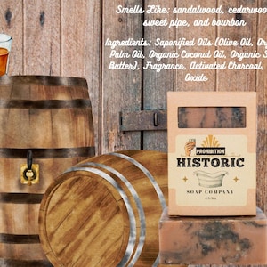 May include: A bar of soap and its packaging from the Prohibition Historic Soap Company. The packaging is tan with the company name and logo. The soap is brown with dark gray marbling. A glass of amber liquid sits atop a wooden barrel.