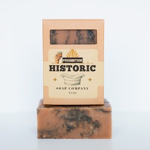 May include: A bar of soap in a cardboard box. The soap is a light pink color with black swirls. The box is brown with a label that says "Prohibition Historic Soap Company 4.5 oz".