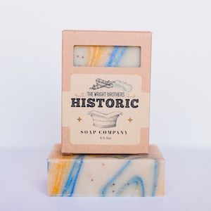 May include: A bar of soap with a marbled design in shades of white, yellow, and blue, with a matching box. The box features the text "THE WRIGHT BROTHERS HISTORIC SOAP COMPANY" and a graphic of an airplane and a tub.