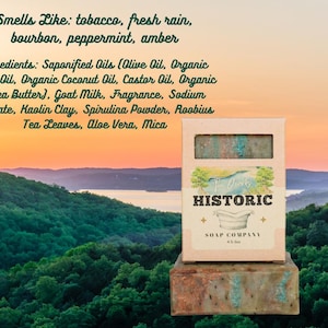 May include: A bar of soap in a cardboard box with the text "Historic Soap Company." The soap is green and brown, with a view of a lake and mountains in the background. The text on the box says the soap smells like tobacco, fresh rain, bourbon, peppermint, and amber.