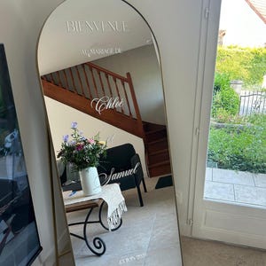 Peut inclure: Un miroir sur pied arqué avec un cadre fin doré portant l'inscription en vinyle blanc BIENVENUE AU MARIAGE DE Chloé et Samuel 13 JUIN 2026. Le miroir reflète un intérieur avec un escalier en bois, une chaise et un bouquet de fleurs.