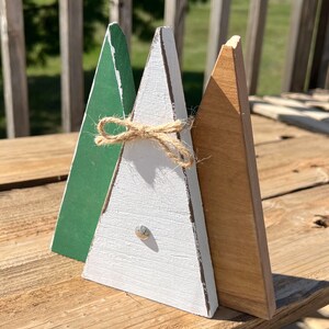 Puede incluir: Tres adornos de árbol de Navidad de madera, uno verde, uno blanco y uno marrón. El árbol blanco tiene un lazo de cuerda y una cabeza de clavo plateada.