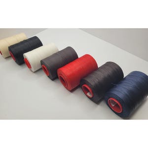 May include: A selection of thread spools in a range of colours: cream, black, white, grey, red, brown, and navy. Each spool has a red core. The tightly wound threads suggest they are suitable for sewing and crafting projects.