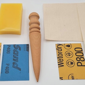 May include: A collection of leather crafting tools and supplies, including a wooden edge beveler, sandpaper, a yellow wax block, and canvas pieces. The sandpaper is labeled P600, P400, and P800. The tools are arranged on a white surface.