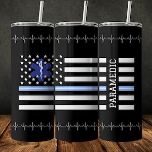 Könnte beinhalten: Drei Edelstahlbecher mit einem schwarz-weißen amerikanischen Flaggenmuster. Der mittlere Becher zeigt ein blaues medizinisches Symbol mit einer Schlange und einem Stab. Der rechte Becher hat das Wort "PARAMEDIC" vertikal gedruckt. Alle drei Becher haben eine dünne blaue Linie über dem Flaggenmuster.