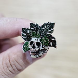 May include: A detailed enamel pin featuring a skull with a leafy green plant design. The skull is white with black details, and the leaves are dark green with lighter green accents. The pin is a unique accessory.