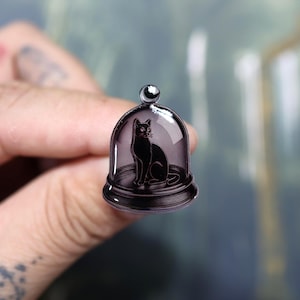 May include: A black cat illustration inside a glass dome. The cat is sitting, with its tail curled. The dome has a small, round top. The base of the dome is a dark, circular shape. The overall design is a dark, gothic style.