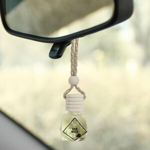 May include: A white and gold car air freshener with a clear glass bottle and a wooden cap. The bottle is filled with a clear liquid and has a label that says "RAIN WATER".
