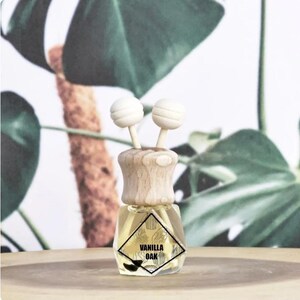 May include: A small, clear glass bottle with a wooden cap and two white, spherical diffusers. The bottle contains a yellow liquid and a label that reads "VANILLA OAK". The background features green leaves and a wooden surface.