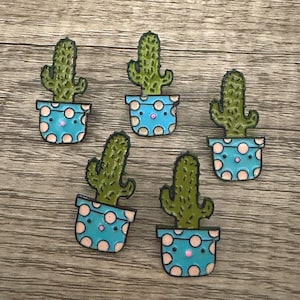 May include: Five cactus-shaped pins. Each pin features a green cactus in a blue pot with white polka dots. The pins have a black outline and are arranged on a wooden surface.