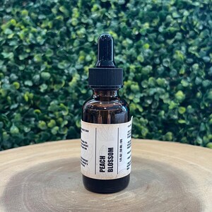 May include: A brown glass bottle with a black dropper top, labeled "Peach Blossom" with a white label. The bottle contains 1 fl oz / 30 ml of oil. The background is a green leafy wall, and the bottle sits on a wooden surface.