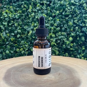 May include: A brown glass bottle with a black dropper top. The label reads "MOROCCAN CASHMERE" and "1 fl oz | 30 ml oil". The bottle is on a wooden surface with a green leafy background.