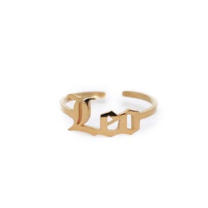 May include: A gold-colored ring with the word "Leo" in an Old English font. The ring is open at the back and sits on a white background. The ring is a jewelry item.