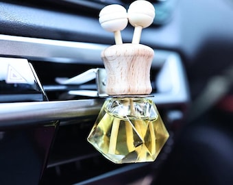 Könnte beinhalten: Ein rautenförmiger, gläserner Autolufterfrischer mit einem Holzdeckel und zwei weißen Diffusorsticks. Der Lufterfrischer ist an einem Lüftungsschlitz im Auto befestigt.