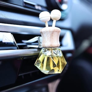 May include: A diamond-shaped glass car air freshener with a wooden top and two white diffuser sticks. The air freshener is attached to a car's air vent.