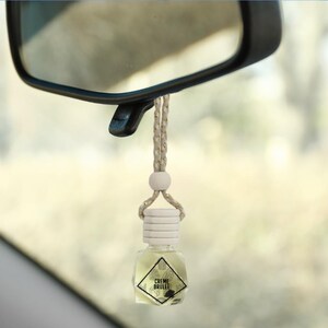 May include: A white and gold car air freshener with a clear glass bottle and a wooden cap. The bottle is filled with a light yellow liquid and has a label that reads "CREME BRULEE".