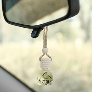 May include: A car air freshener with a small, clear, square-shaped bottle filled with a light yellow liquid. The bottle has a white cap and is suspended by a beige and gold string. The label reads "AMBER DRIFTWOOD". The freshener hangs from a car's rearview mirror.