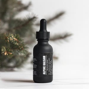 May include: A black glass bottle with a dropper top, labeled "Alpine Balsam" with fragrance notes listed. The bottle is sitting on a white surface with a green leafy branch in the background.