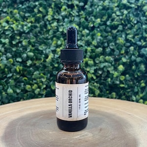 May include: A brown glass bottle of Vanilla Orchid oil with a black dropper and cap. The label reads "Vanilla Orchid" and "1 fl oz / 30 ml Oil". The bottle sits on a wooden surface against a green leafy background.