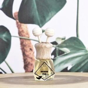 May include: A small, clear glass bottle with a wooden cap and two white ball-shaped diffusers. The bottle contains a yellow liquid and a label that reads "Orchard Pear". The background features green leaves.
