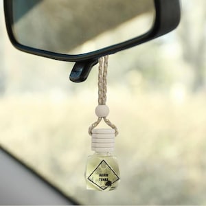 May include: A small, clear glass car air freshener with a diamond-shaped label that reads "WARM TONKA." The freshener hangs from a rearview mirror by a beige and gold string. The liquid inside is light yellow.