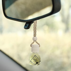 May include: A clear glass bottle with a white lid and a gold string hanging from a rearview mirror. The bottle has a diamond-shaped design and the text "ROSE + OUD" printed on the label.