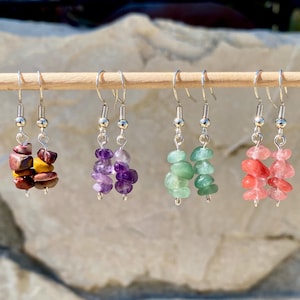 Puede incluir: Pendientes colgantes con ganchos plateados. Cada par presenta una pila de pequeñas piedras preciosas de forma irregular en varios colores: rojo, amarillo, morado, verde y rosa. Los pendientes se exhiben en una varilla de madera.
