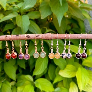 以下が含まれることがあります： 緑の葉を背景に、木の棒に飾られた宝石のイヤリングのコレクション。 イヤリングは、赤、紫、緑、黄色、白と黒の斑点など、さまざまな色の石が特徴で、すべてワイヤーで包まれ、金または銀のフックから吊り下げられています。