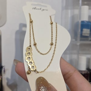 Może przedstawiać: Złota bransoletka na kostkę z akcentami monet i koralików. Bransoletka na kostkę posiada szereg małych, okrągłych koralików i uroków w kształcie monet. Jest prezentowana na białej karcie z napisem "thank you".