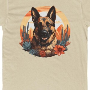 May include: Beige t-shirt with a German Shepherd dog design set against a desert sunset. The illustration includes cacti, succulents, and orange flowers. The dog's portrait is detailed, with a happy expression.
