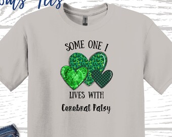 May include: A light gray t-shirt with a green shamrock design and the text "Some One I Lives With Cerebral Palsy". The shirt is shown with a pair of blue jeans, sunglasses, and white sneakers.