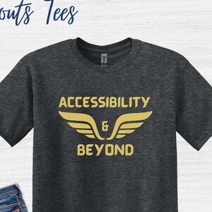 May include: A dark heather grey t-shirt with the text "Accessibility & Beyond" in gold lettering with wings above the text. The shirt is paired with blue jeans and black high-top sneakers.