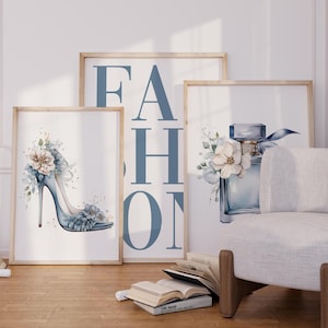 May include: Three framed watercolour art prints. One depicts a high-heeled shoe adorned with flowers, another a perfume bottle with floral accents, and the third displays the word "FASHION" in large blue letters. The prints are displayed in a room with a chair and books.