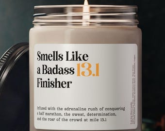 May include: A lit candle in a glass jar with a black lid. The label reads "Smells Like a Badass 13.1 Finisher." The text below describes the scent as "infused with the adrenaline rush of conquering a half marathon."