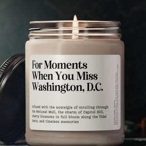 May include: A lit candle in a glass jar with a silver lid and a black lid next to it. The label reads "For Moments When You Miss Washington, D.C." with text below describing the scent and theme.