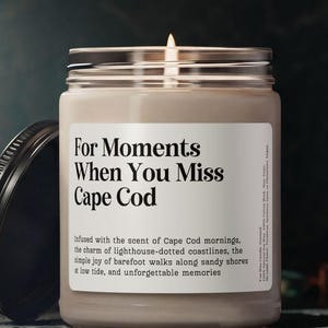 May include: A lit candle in a glass jar with a silver lid and a black lid next to it. The label reads "For Moments When You Miss Cape Cod". The candle is beige and the background is dark.