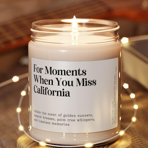 May include: A lit candle in a clear glass jar with a white label that reads "For Moments When You Miss California." The candle sits on a round wooden coaster, surrounded by string lights. A guitar and a book are in the background.