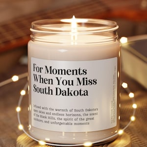 May include: A lit candle in a glass jar with a label that reads "For Moments When You Miss South Dakota." The candle sits on a small wooden coaster, surrounded by string lights. A guitar and a book are in the background.