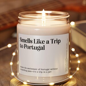 May include: A lit candle in a glass jar with the text "Smells Like a Trip to Portugal" on a white label. The candle sits on a round wooden coaster, with string lights and a guitar in the background.