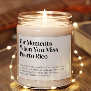 May include: A lit candle in a glass jar with a label that reads "For Moments When You Miss Puerto Rico." The candle sits on a wooden coaster, surrounded by string lights. A guitar and a book are in the background.