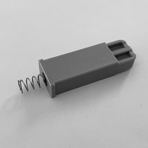 May include: A gray plastic component with a metal spring attached. The rectangular piece has a grooved end. The spring is coiled and extends from one side of the plastic piece. The image is in grayscale.