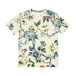 May include: A cream-coloured t-shirt with a floral design in blue, green, and red. The pattern features stylised flowers, leaves, and branches, creating a botanical theme. The shirt has short sleeves and a crew neck.