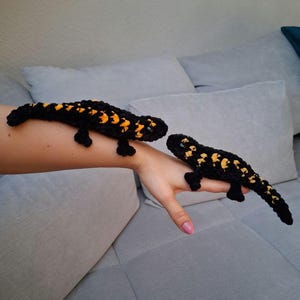 Peut inclure: Deux peluches de salamandres au crochet, l'une noire avec des marques orange et l'autre noire avec des marques jaunes, sont posées sur le bras d'une personne.