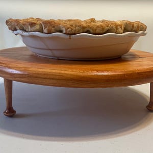 May include: A wooden pie stand with three turned legs, supporting a pie in a white ceramic pie dish.