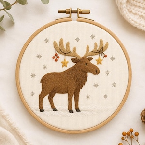 May include: A circular wooden embroidery hoop containing a cream-coloured fabric panel with a hand-stitched brown moose. The moose features golden yellow stars and red berries hanging from its antlers, surrounded by small grey embroidered snowflakes.