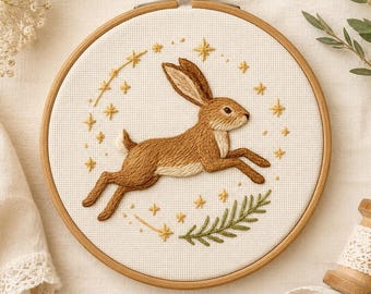 May include: A circular wooden embroidery hoop containing white fabric with a hand-stitched brown rabbit leaping through the air. The rabbit is surrounded by embroidered yellow stars and a small green leafy branch, all centered within the hoop frame.