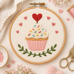 Può includere: Un telaio da ricamo rotondo in legno contenente un pannello di tessuto bianco con un disegno di cupcake ricamato a mano. Il cupcake presenta una base plissettata rosa, una torta color marrone chiaro, glassa bianca con zuccherini colorati e un cuore.